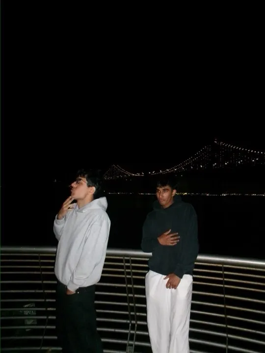Two friends at a railing at night, bridge lights behind them
